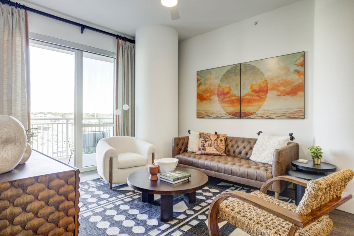 Modern living room with a leather sofa, round table, abstract wall art, and large window with curtain.