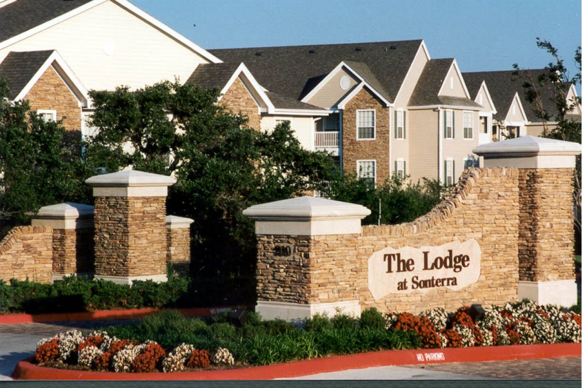 Stone entrance sign for The Lodge at Sonterra apartments, with landscaped flowers and multi-story buildings behind.