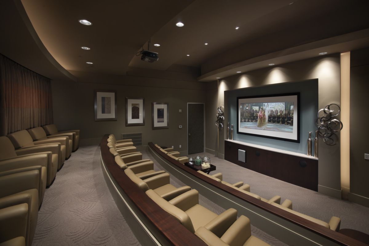 Modern home theater room with curved rows of beige seats facing a large screen displaying a movie.