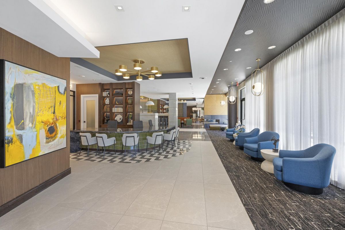 Modern hotel lobby with blue chairs, large artwork, and a check-in desk beneath a stylish light fixture.
