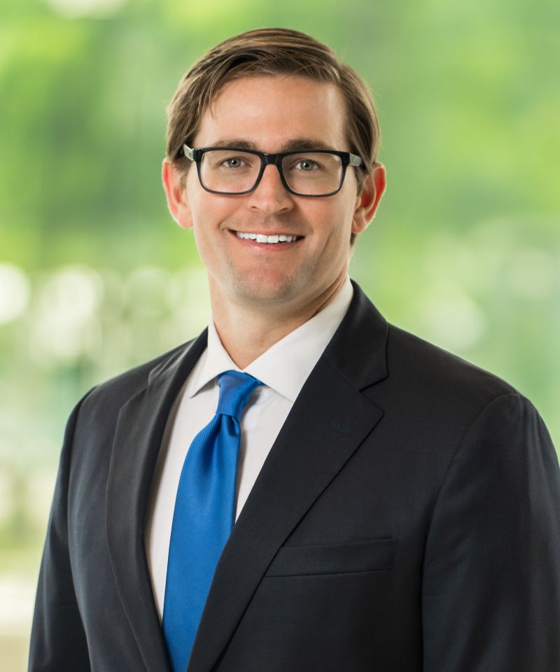 Smiling man in a suit and blue tie with glasses, standing in front of a blurred green background.