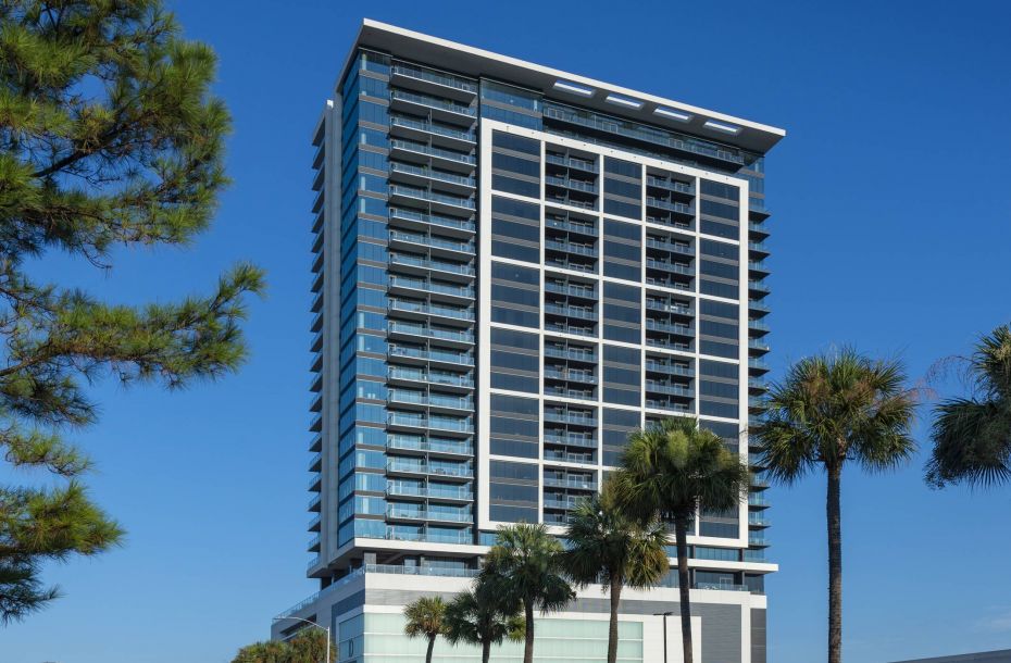Hanover Company, The Driscoll at River Oaks Tall modern building with glass facade, surrounded by palm trees, under a clear blue sky.