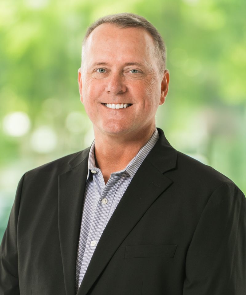 Smiling man in a black blazer and collared shirt standing in front of a blurred green outdoor background.