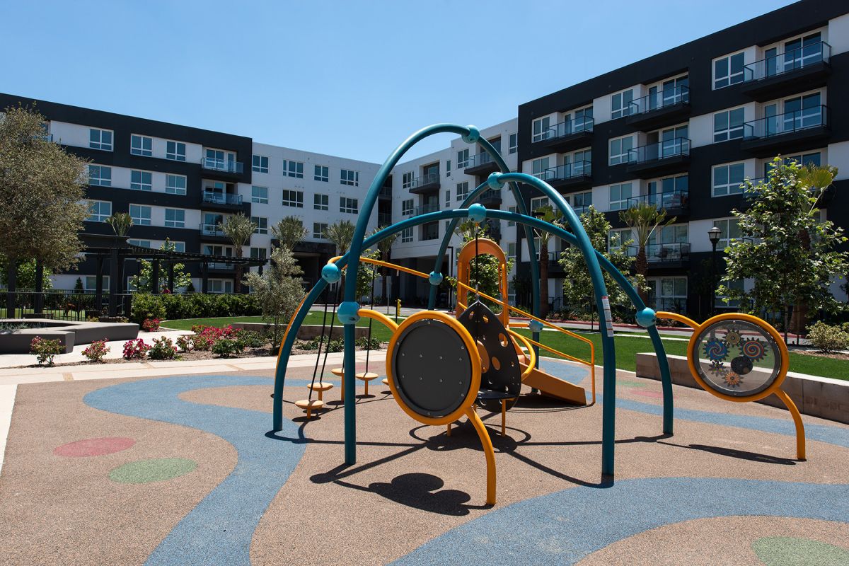 Colorful modern playground with climbing equipment, set in front of multi-story apartment buildings on a sunny day.