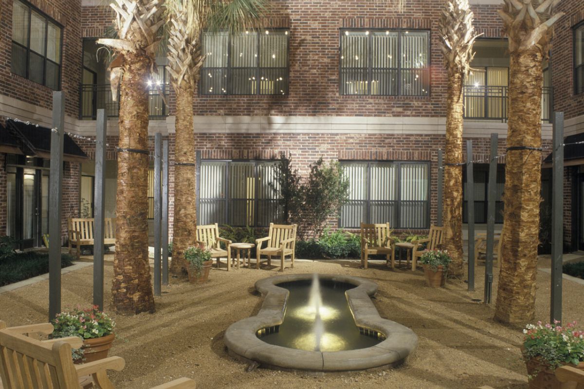 Night courtyard with palm trees, wooden benches, a round fountain, and string lights, surrounded by brick buildings.