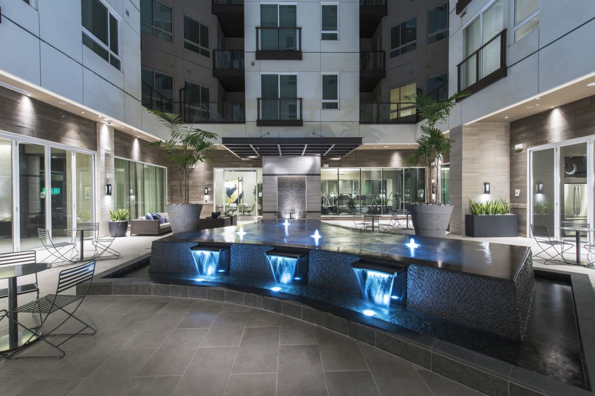 Modern apartment courtyard with illuminated water fountains, patio seating, and large potted plants at night.