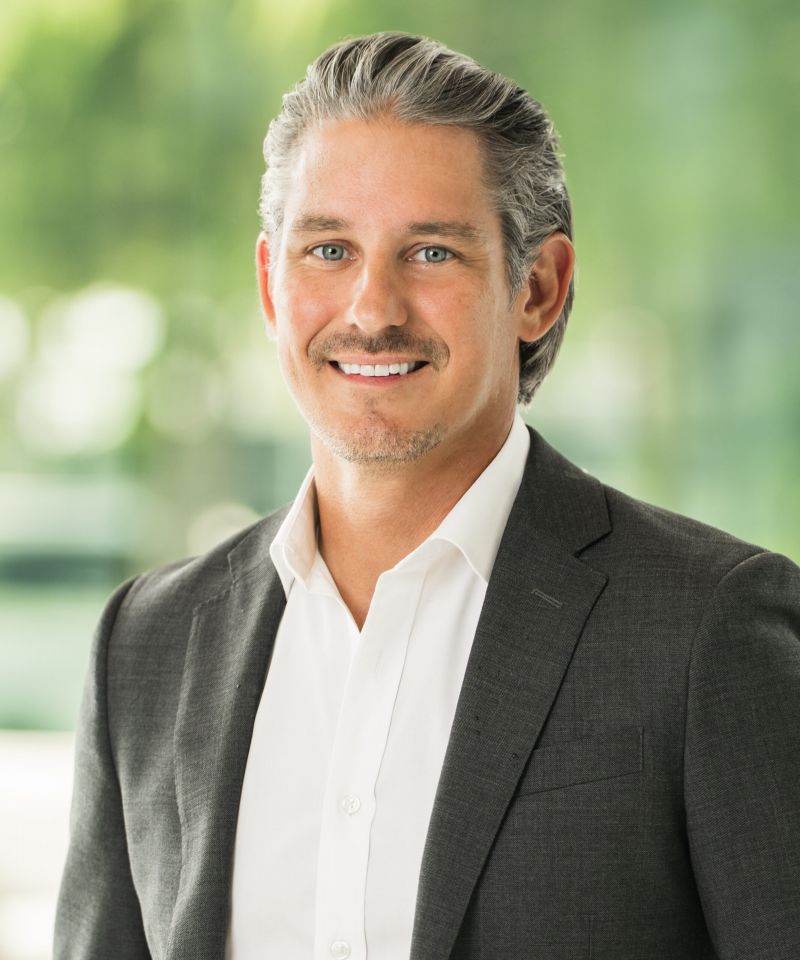 Smiling man in a dark suit and white shirt with blurred green background.