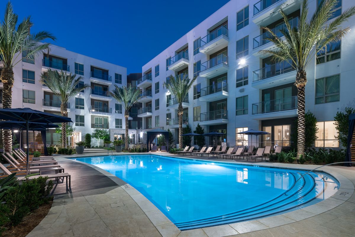Modern apartment complex at night with a lit outdoor pool, lounge chairs, palm trees, and balconies.