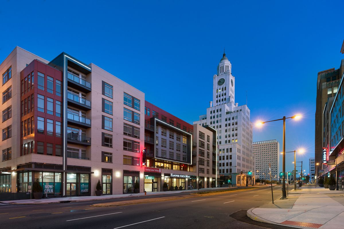 City street at dusk with modern apartments, a clock tower, and streetlights illuminating empty sidewalks.