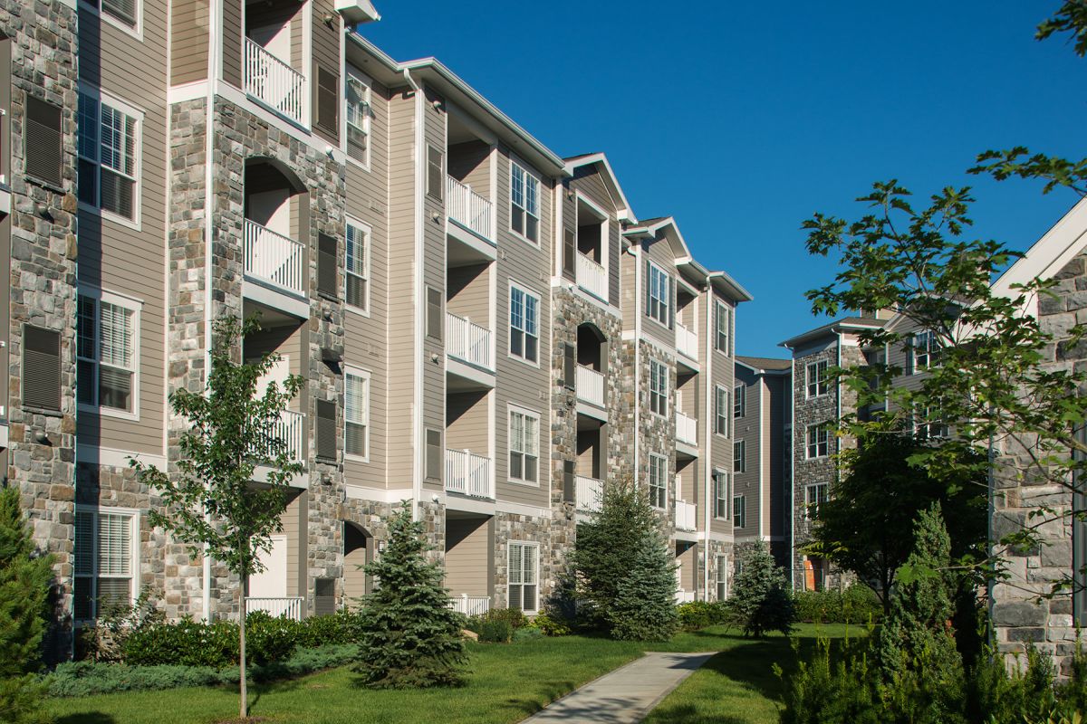 Curved sidewalk between modern apartment buildings with balconies, landscaped lawns, and trees under a blue sky.
