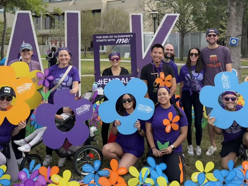 A group of people in purple shirts pose with colorful flower props at a Walk to End Alzheimer’s event.