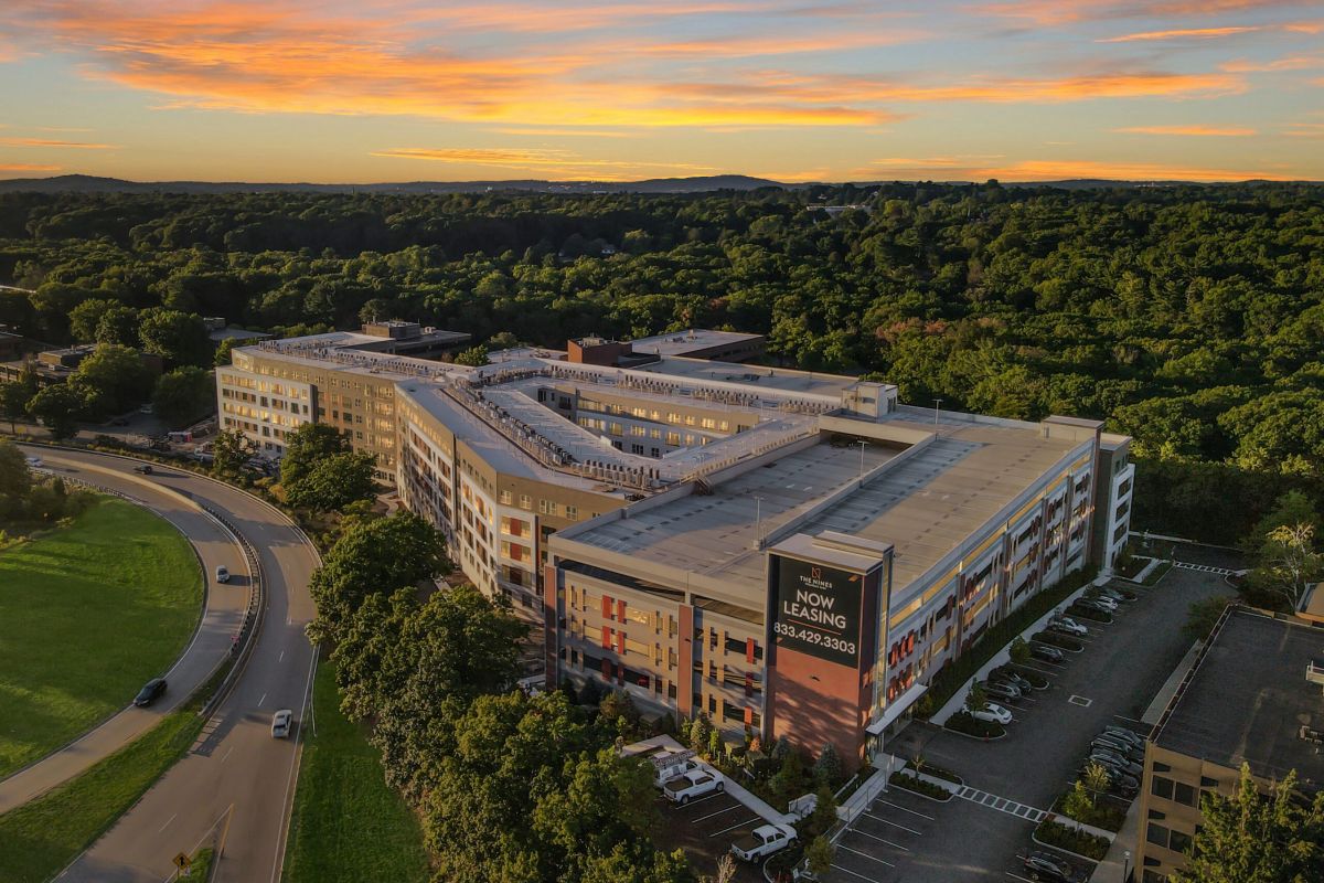 A large apartment complex surrounded by trees at sunset, with a sign reading “Now Leasing.”.