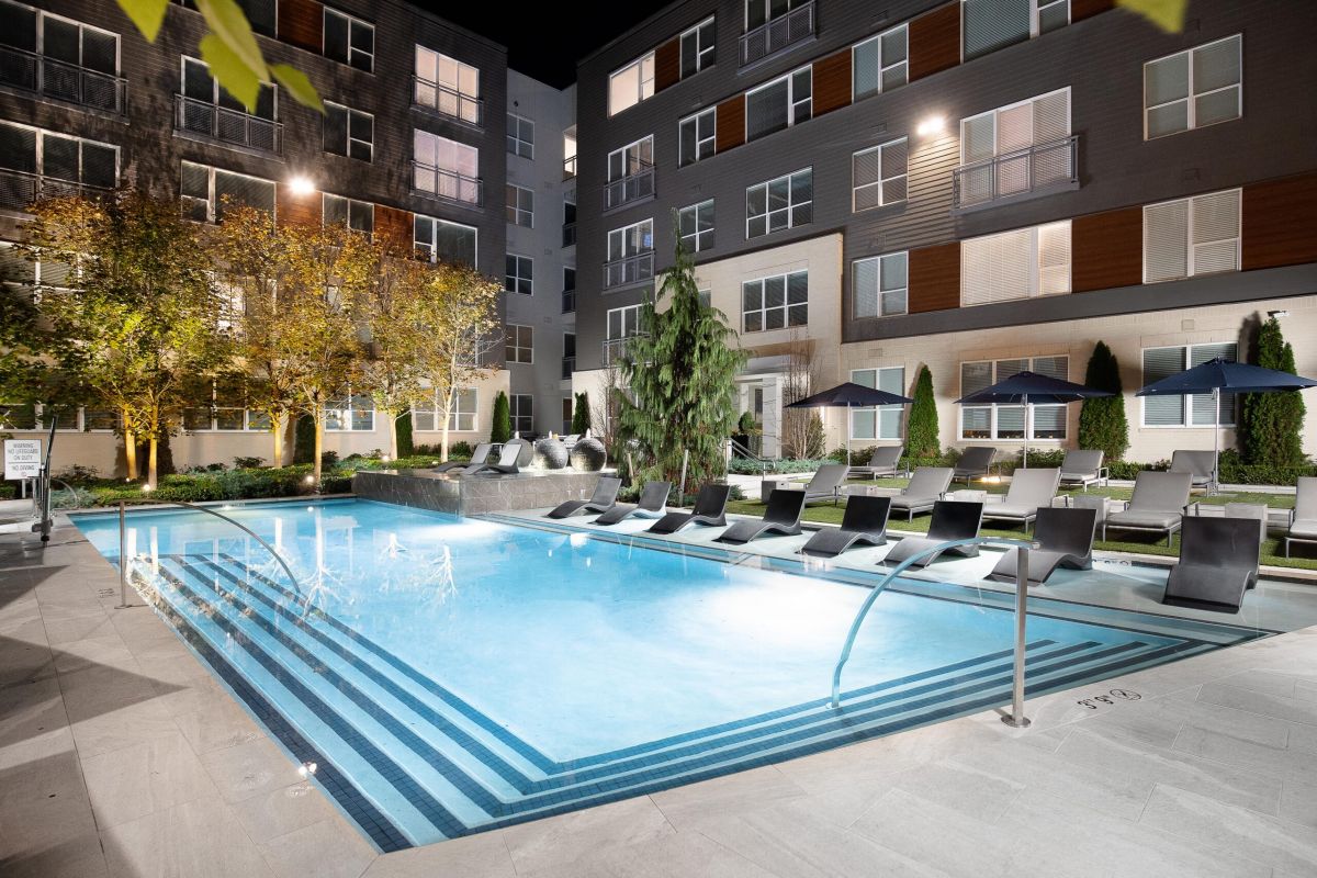 Modern apartment pool at night with lounge chairs, umbrellas, and trees, surrounded by multi-story buildings.