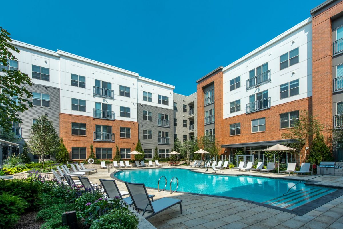 A modern apartment complex with a curved outdoor swimming pool and lounge chairs under a clear blue sky.