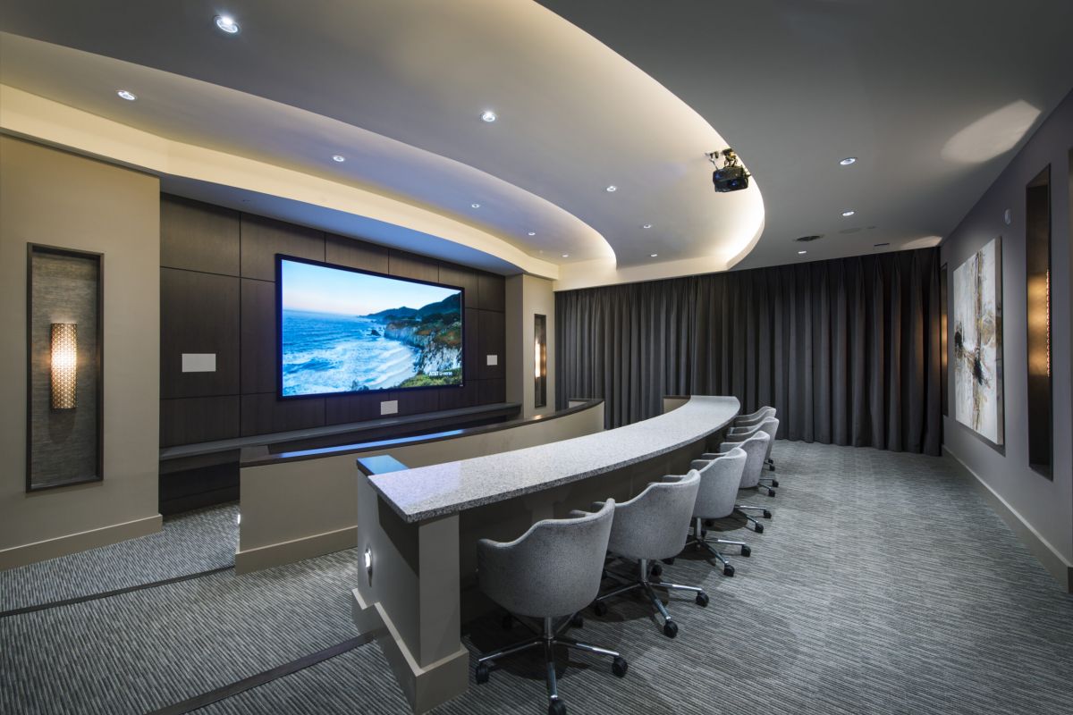 Modern home theater room with curved seating, large screen displaying a beach scene, and gray decor.