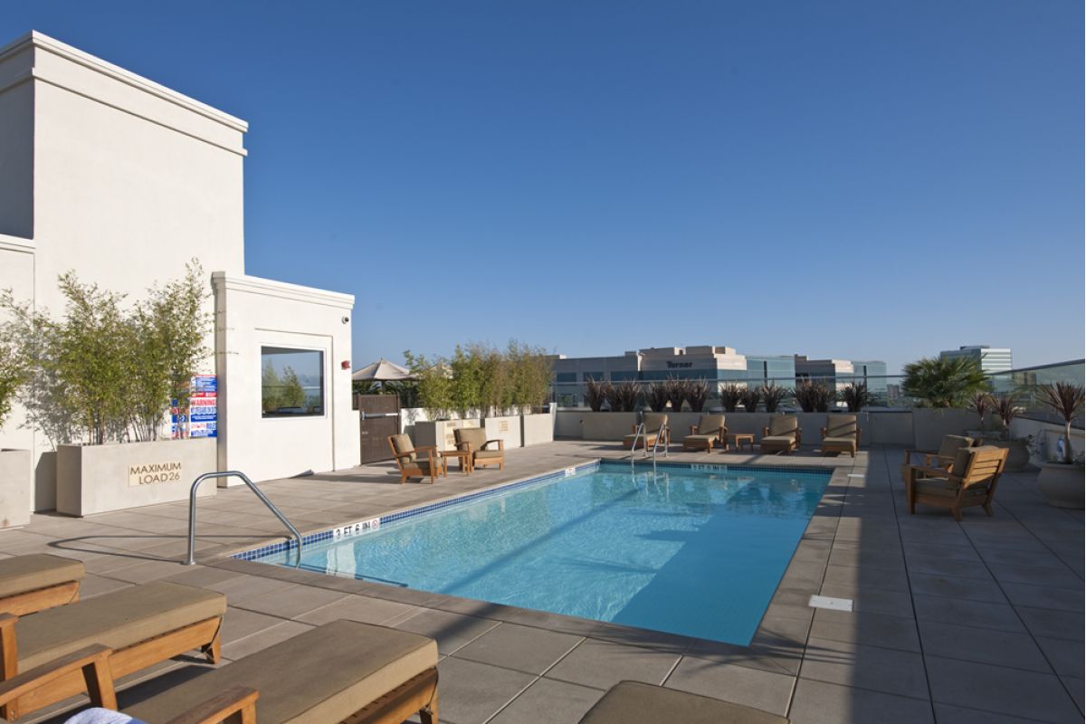Rooftop pool area with lounge chairs, wooden seating, potted plants, and clear blue sky.