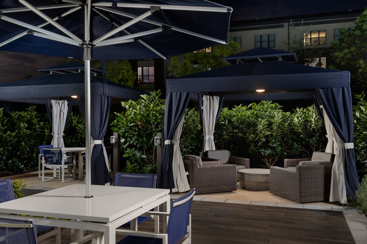 Outdoor lounge area at night with cabanas, wicker chairs, white tables, and large blue umbrellas.