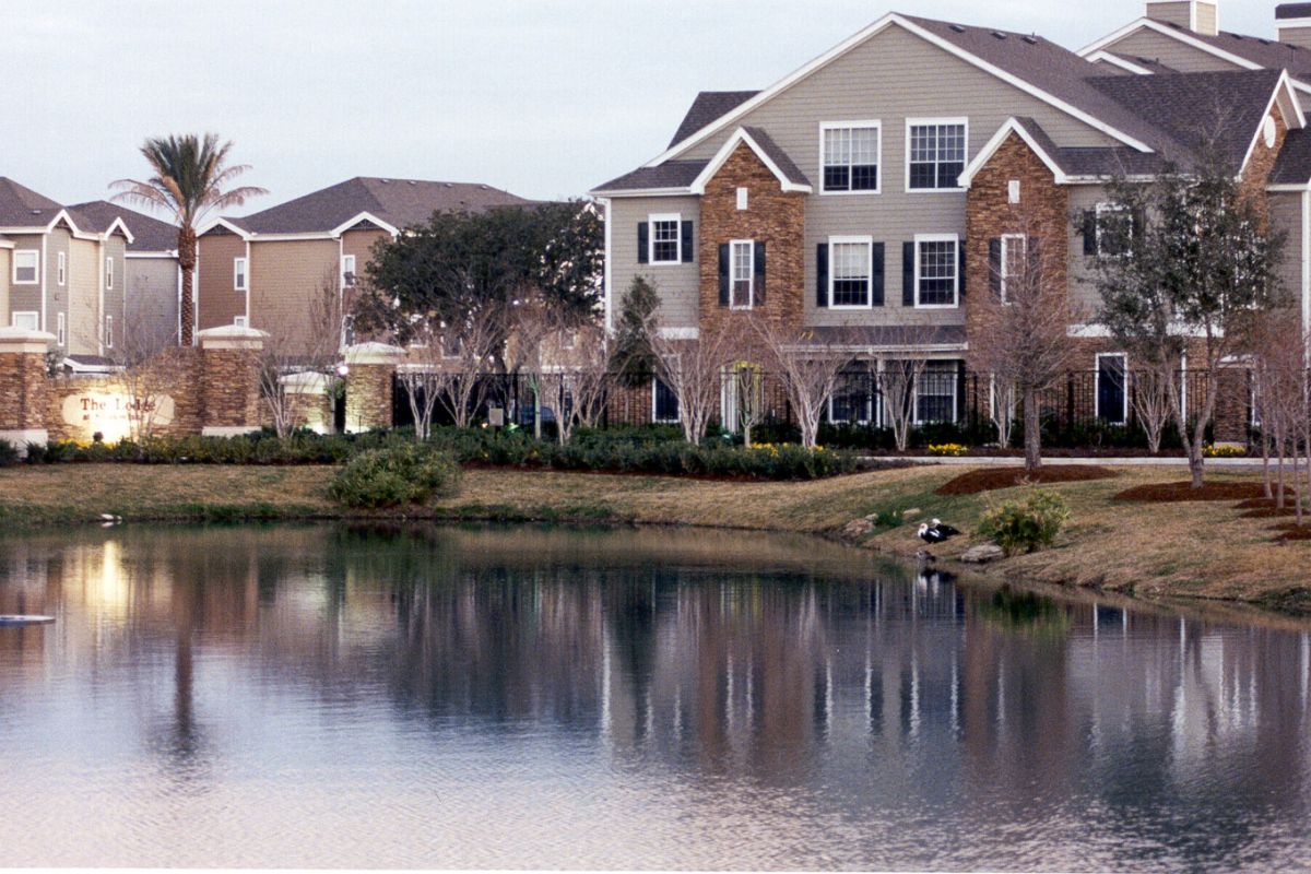 Apartment buildings and trees reflected in a calm pond under an overcast sky.