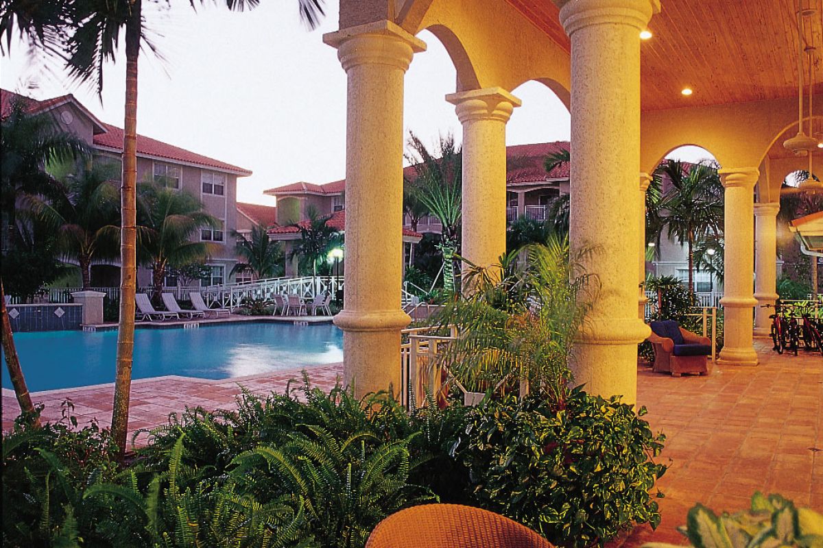 Covered patio with wicker chairs overlooks the Broadwater Galveston pool, framed by palm trees and resort buildings at sunset.