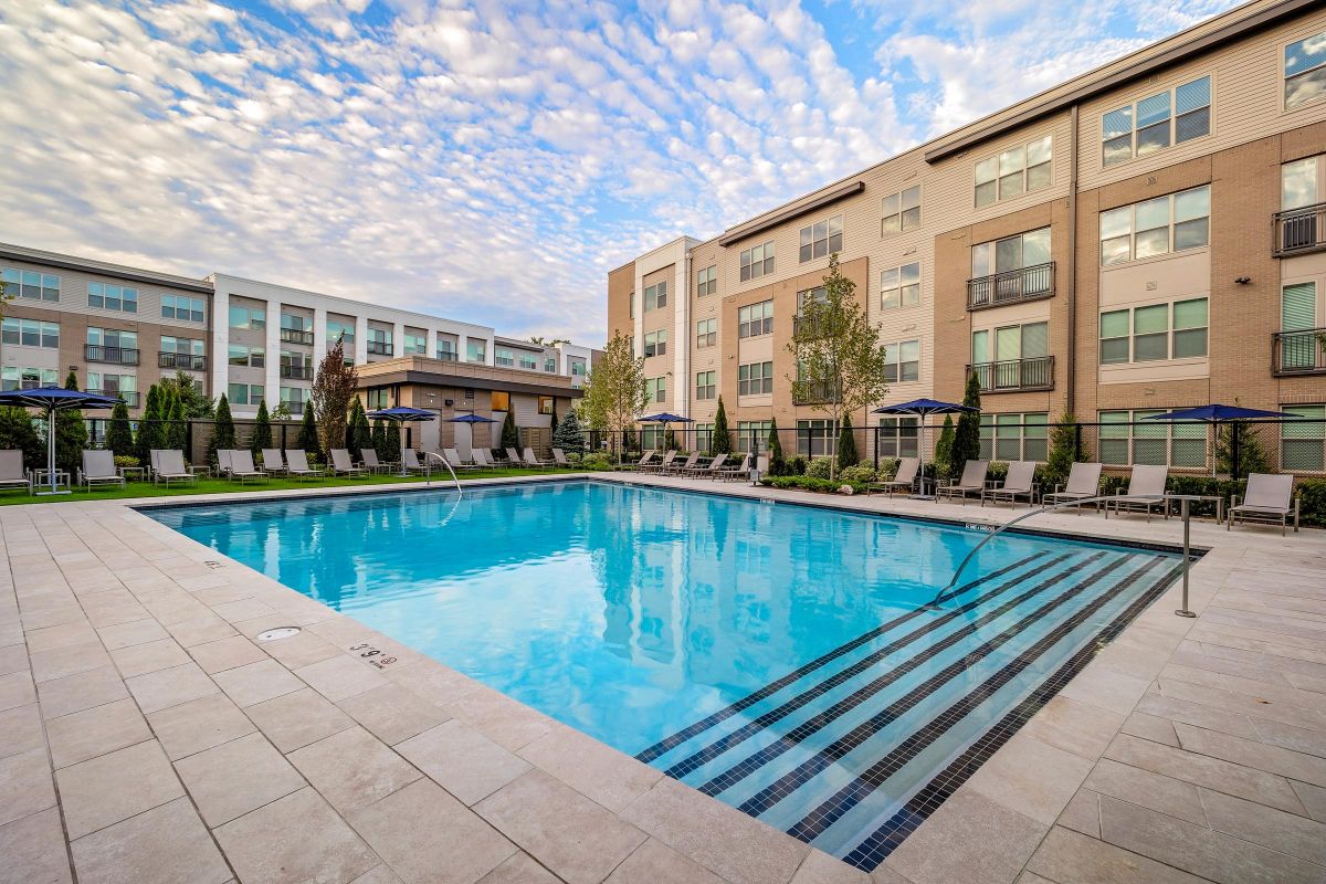 Outdoor swimming pool surrounded by lounge chairs and umbrellas, with modern apartment buildings in the background.