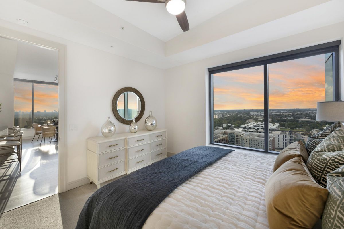 Bedroom with large window showing a city view at sunset, featuring a bed, dresser with round mirror, and neutral decor.