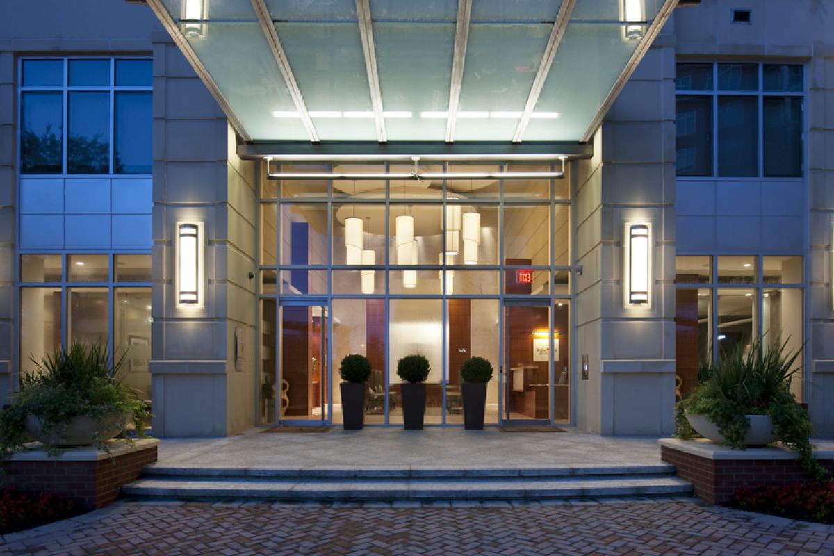 Modern building entrance with glass canopy, potted plants, and illuminated wall lights at night.