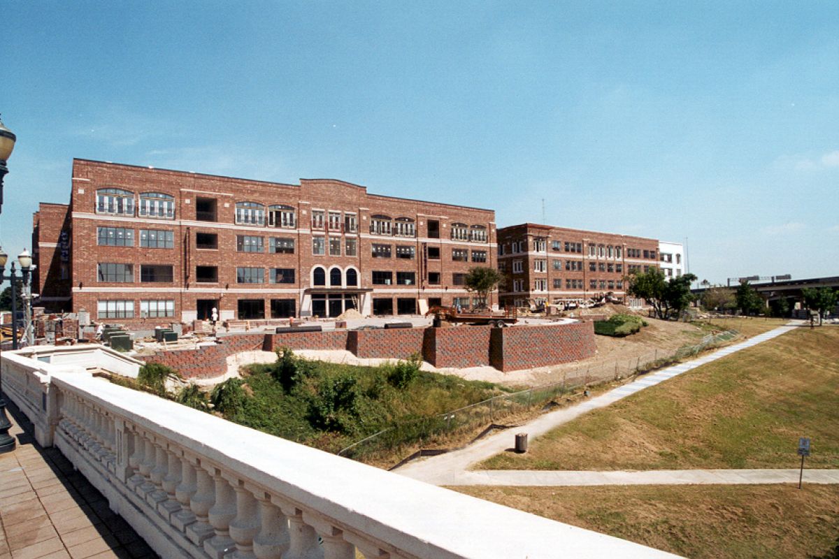 Large red-brick building under construction, seen from across a sunny grassy area and a white railing.