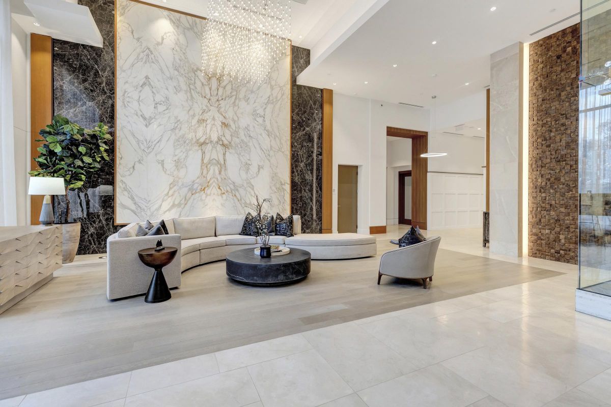 Spacious modern lobby with a curved sofa, marble wall, chandelier, and decorative plants.