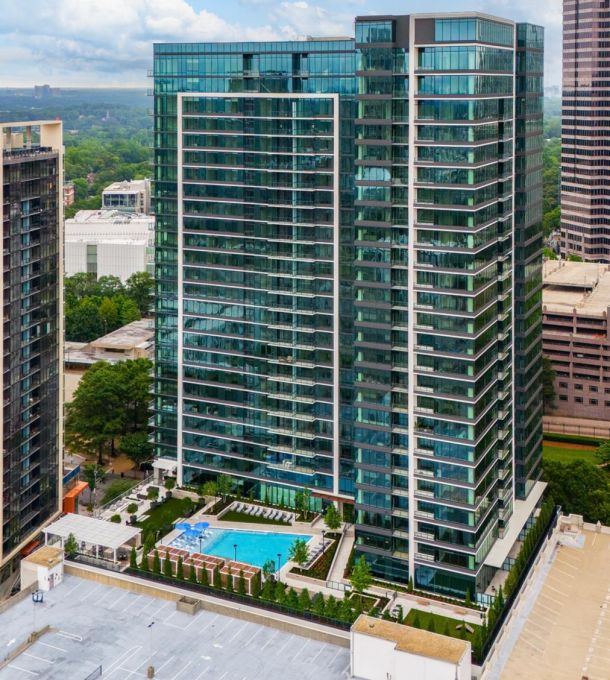 Tall glass apartment building with a pool and trees at its base, surrounded by other high-rise buildings.
