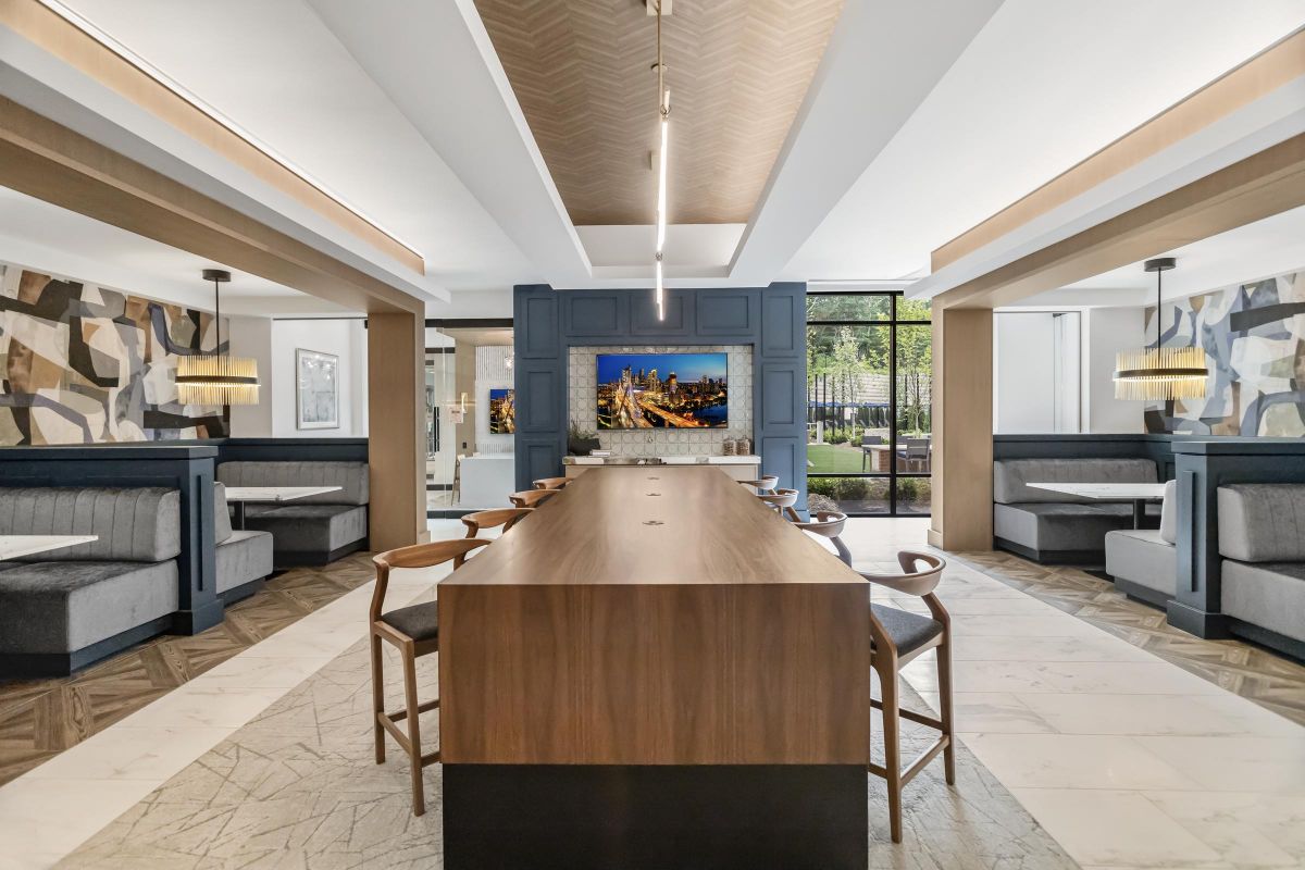 Modern lounge with a long wooden table, surrounding chairs, booth seating, and a wall-mounted TV showing a cityscape.