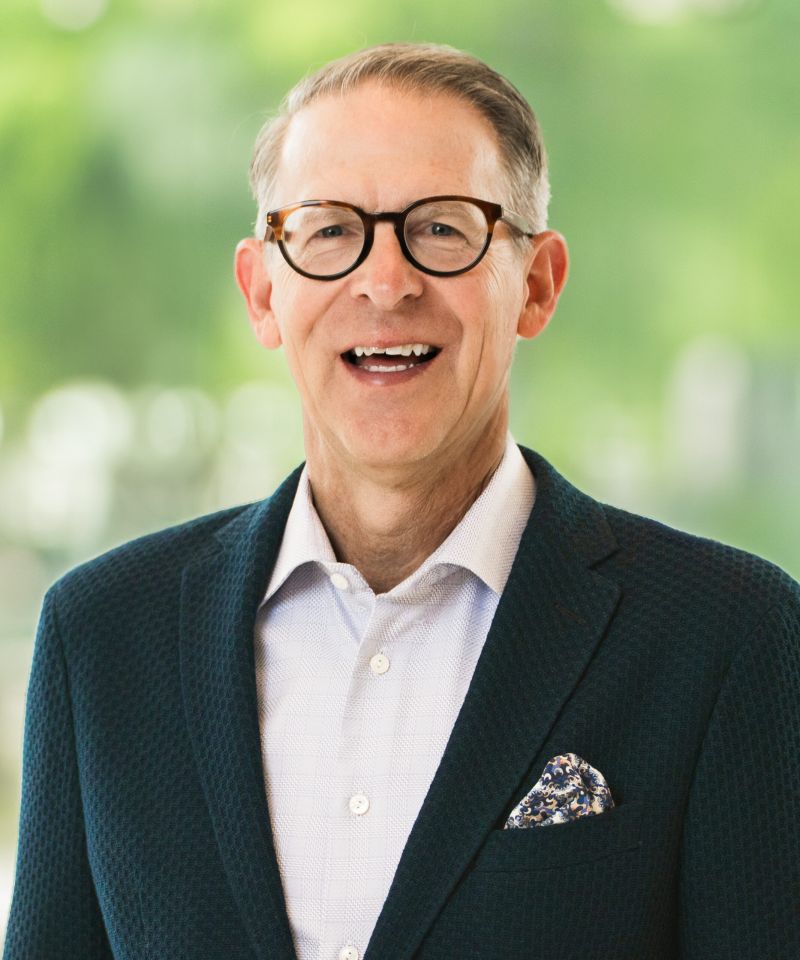 Smiling man with glasses in a blazer and dress shirt, standing in front of a blurred green background.