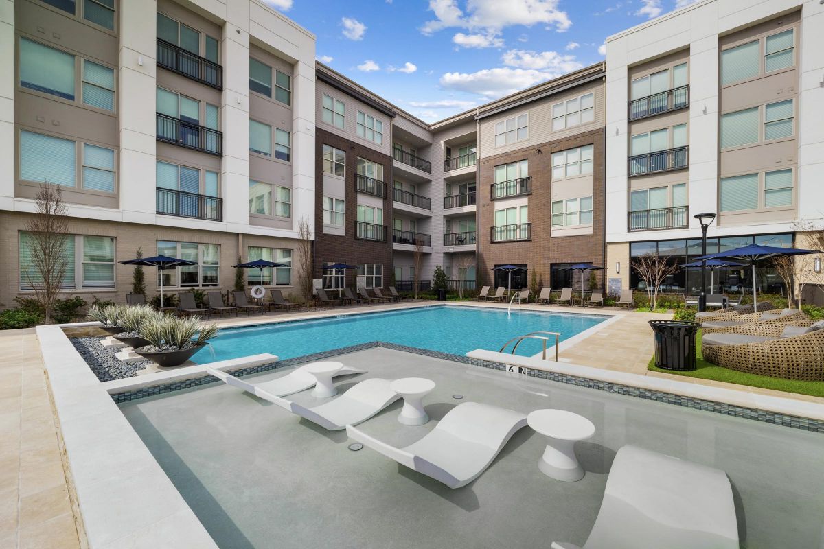 Modern apartment complex with a pool, lounge chairs, and seating area under a partly cloudy sky.