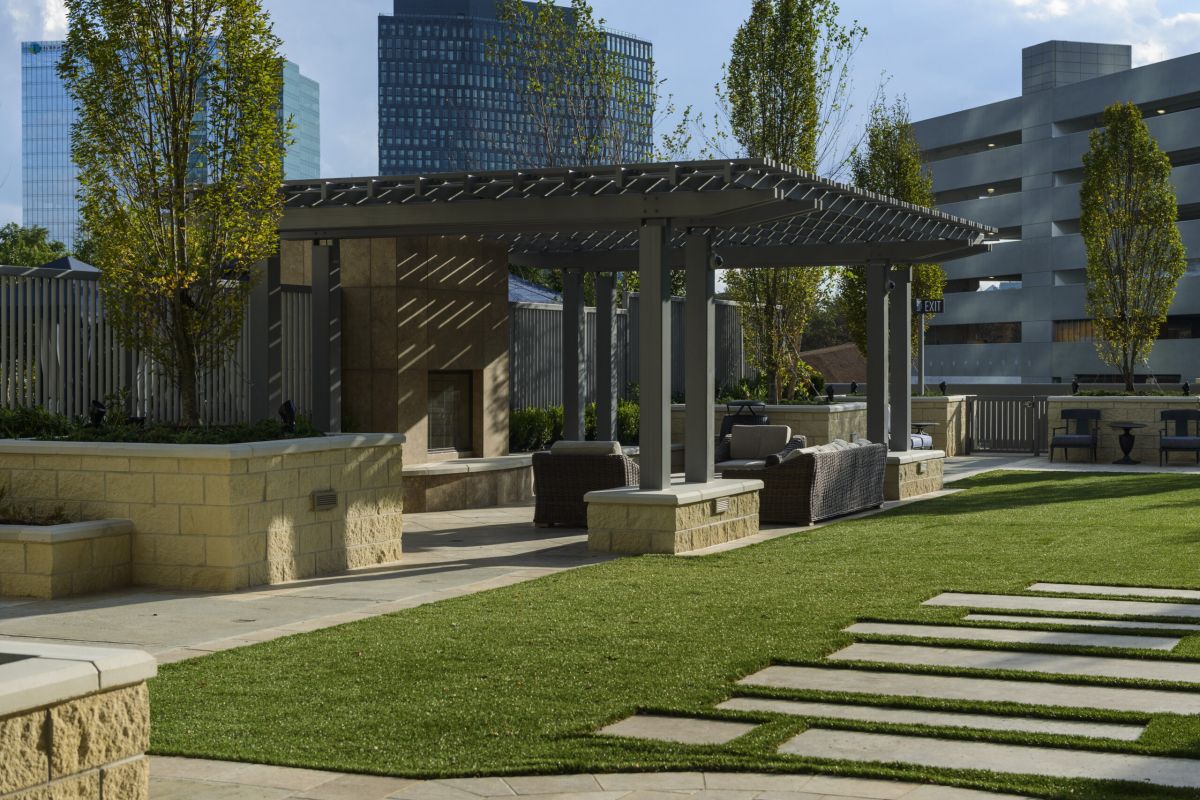 Outdoor patio with pergola, seating area, green lawn, and modern buildings in the background.