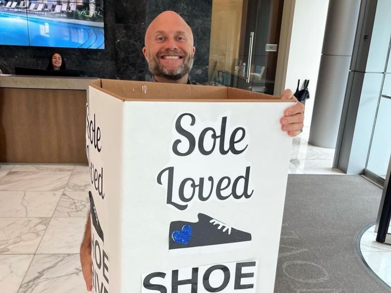 Smiling man holds a large donation box labeled "Sole Loved Shoe" in a modern lobby area.