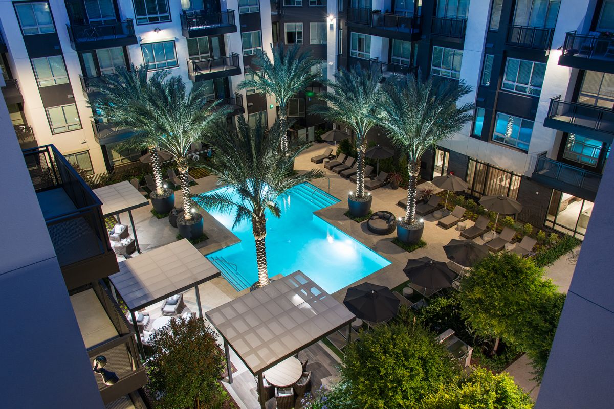 A modern apartment courtyard with a lit swimming pool, palm trees, lounge chairs, and umbrellas at night.