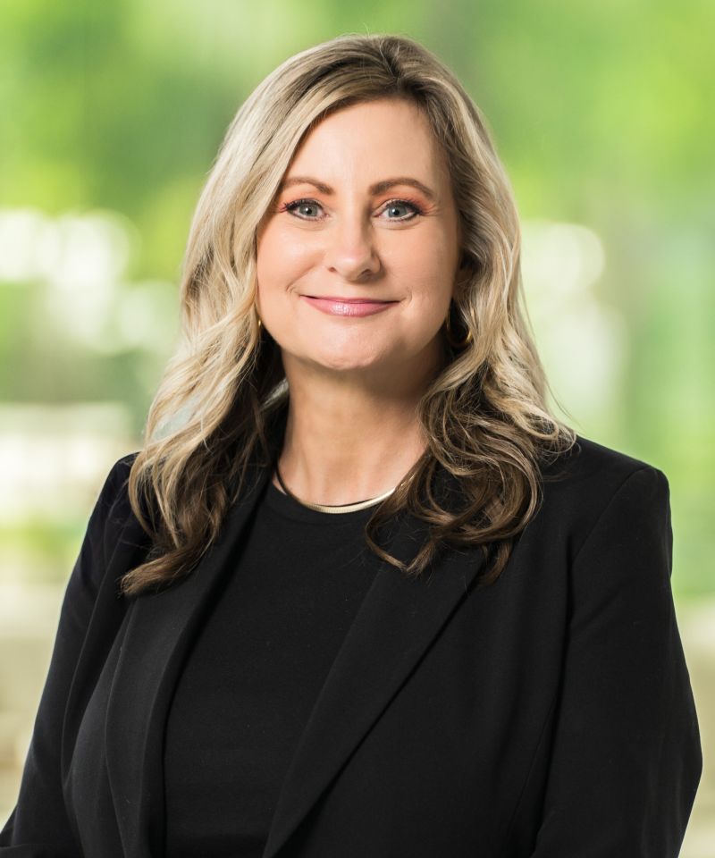 A woman with wavy blonde hair in a black blazer smiles in front of a blurred green background.