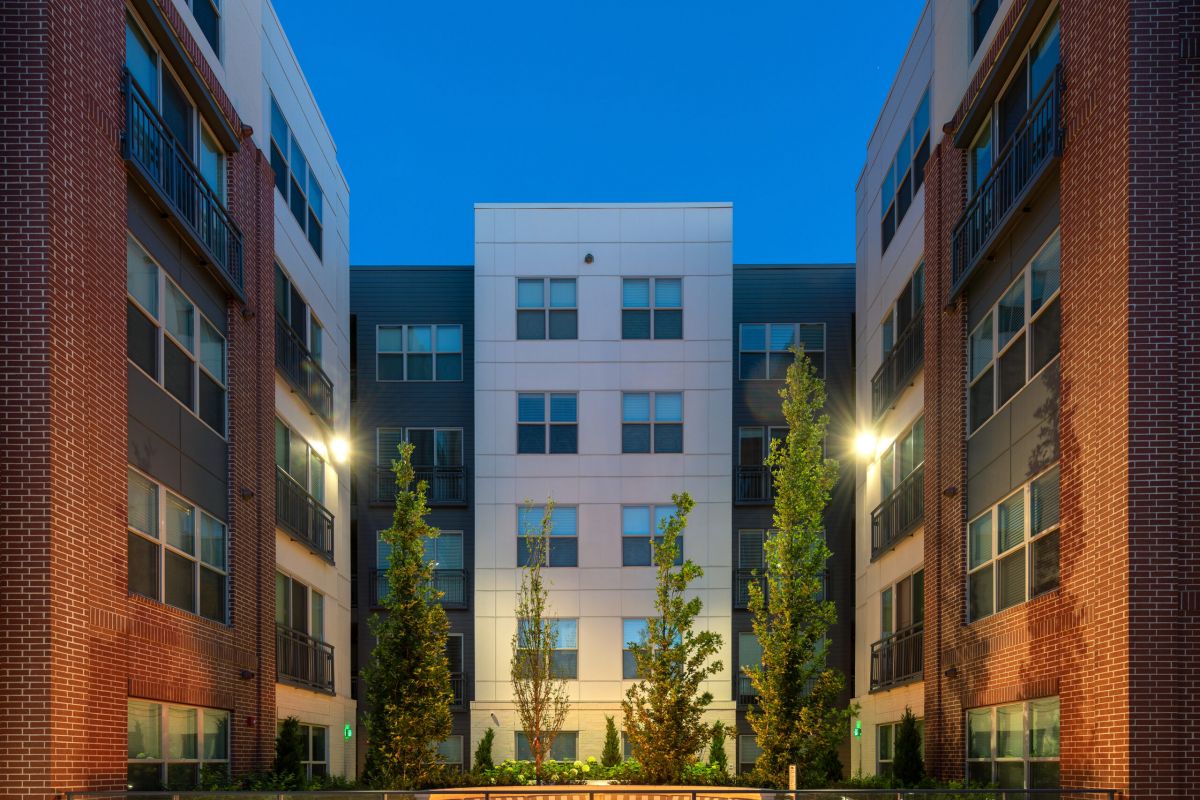 Modern apartment courtyard at dusk with lit benches, trees, and surrounding multi-story buildings.