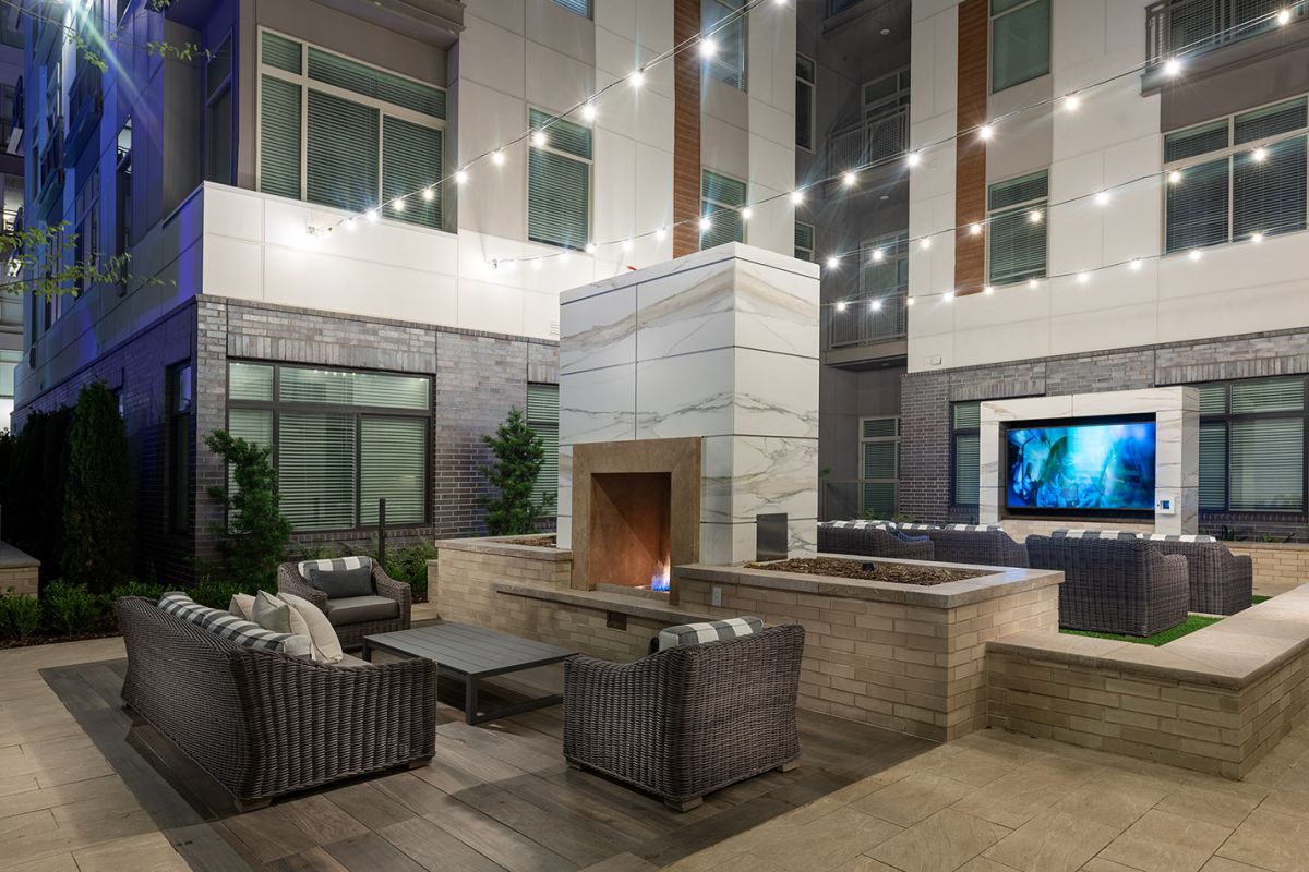 Modern outdoor lounge with wicker seating, a marble fireplace, string lights, and a TV on the wall at night.