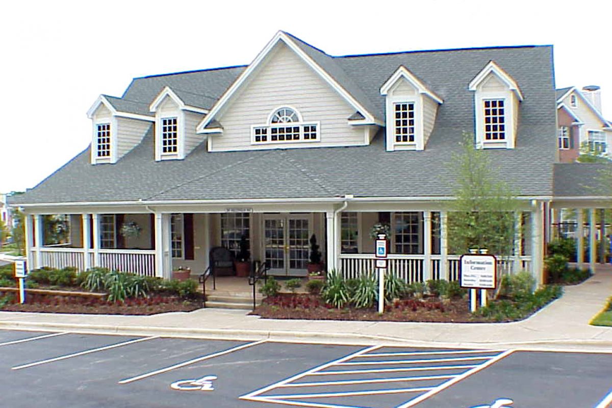 A large, two-story building with a wraparound porch and dormer windows, seen from a parking lot with handicap spaces.