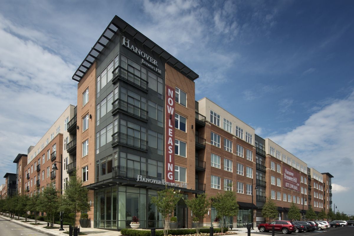 Modern apartment complex with a "Now Leasing" banner, street view, and parked cars under a blue sky.