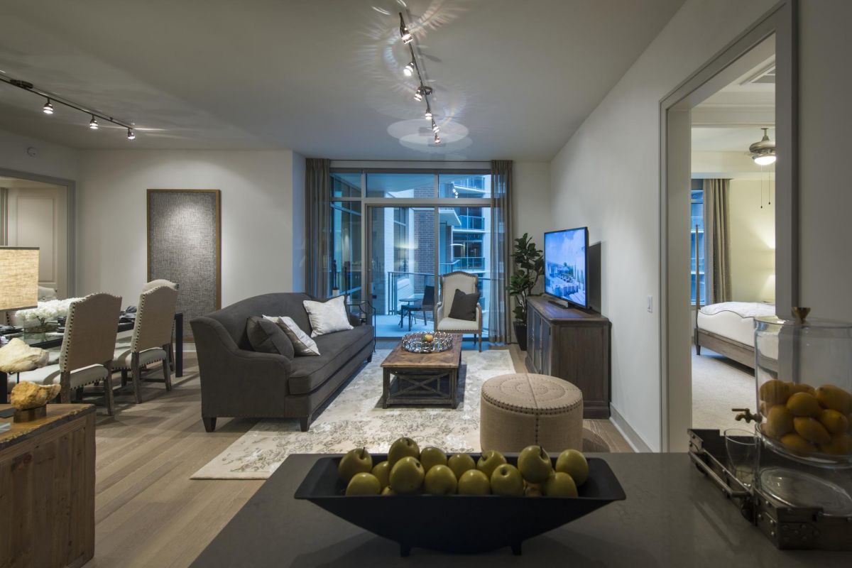 Modern living room with gray sofa, TV, dining area, and fruit bowl on counter; bedroom visible through doorway.