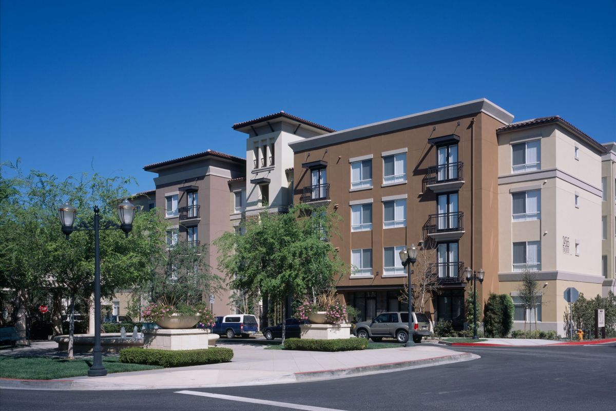 A modern, four-story hotel with beige and brown exterior, trees, and parked cars in front.