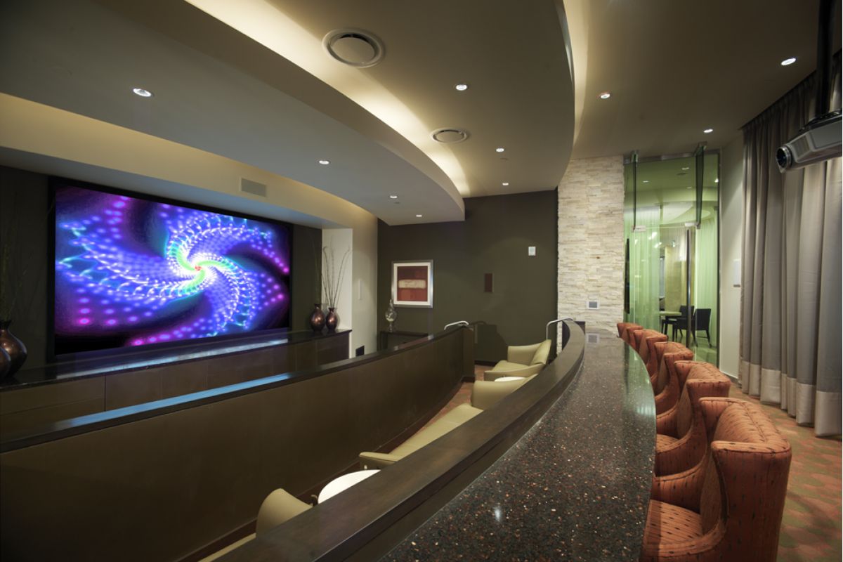 Modern home theater with curved seating, large screen displaying colorful spiral, and bar with orange chairs.