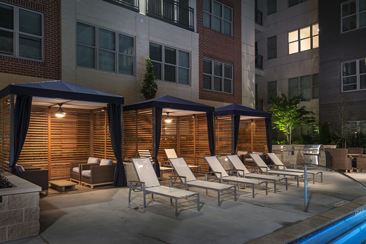 Three poolside cabanas with lounge chairs at night, illuminated by warm lighting next to an apartment building.