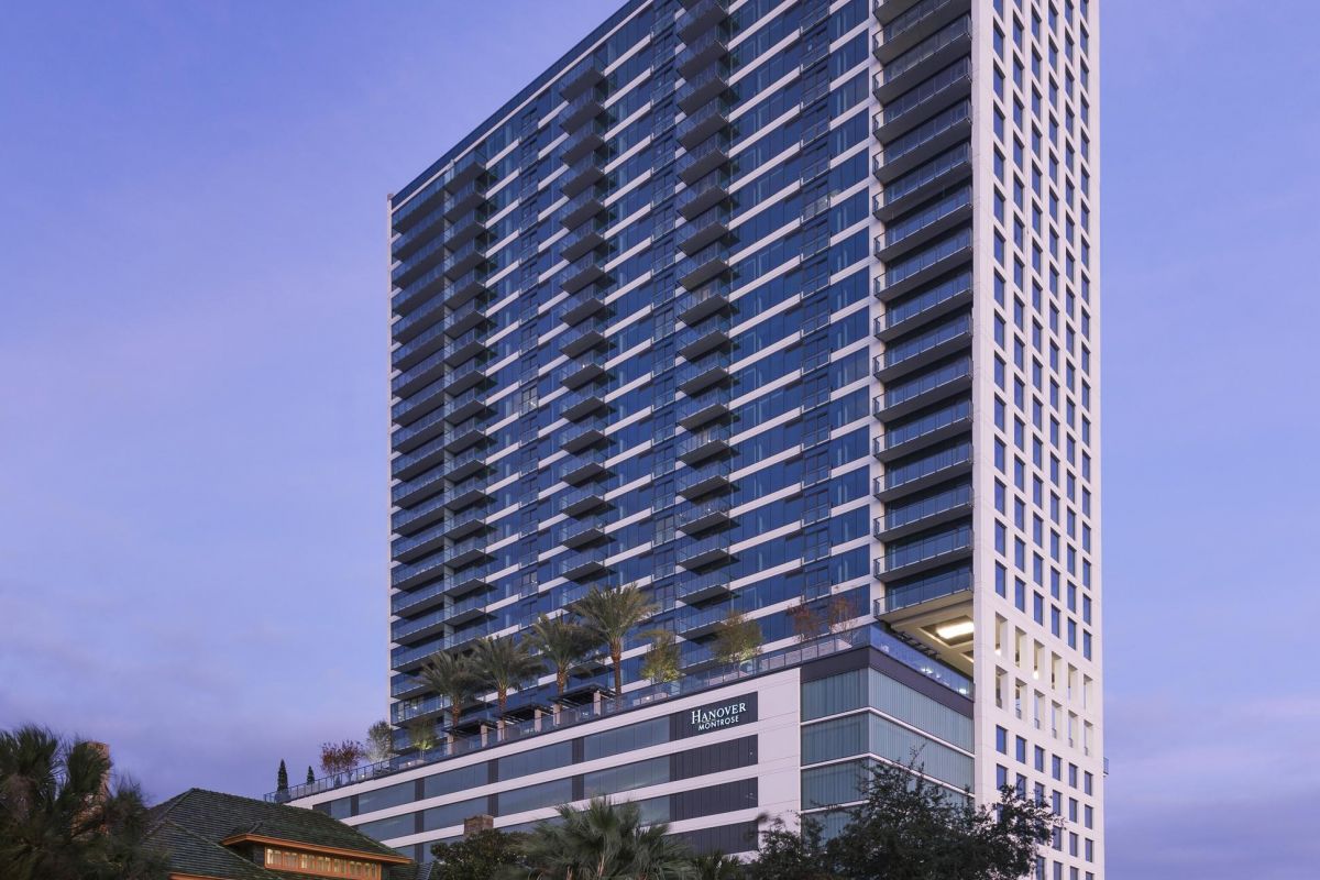 Tall modern building with numerous balconies at dusk, surrounded by trees and smaller structures, under a purple sky.