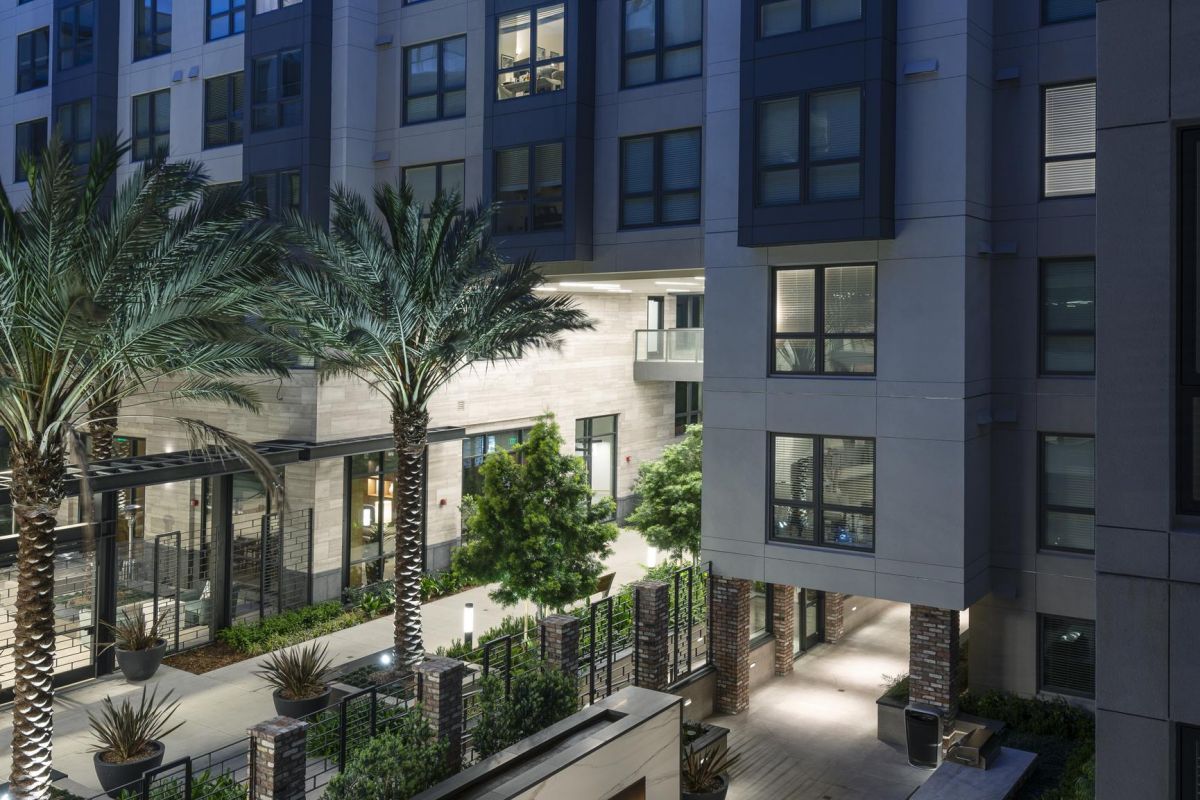 Modern apartment courtyard with palm trees, outdoor seating, and a large fireplace, under a twilight sky.