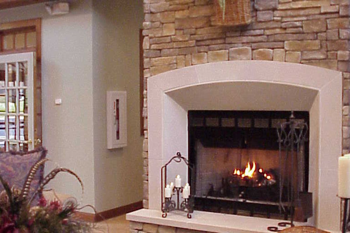 Stone fireplace with a fire burning, floral decor above, cozy chairs, and a wooden table in a living room.