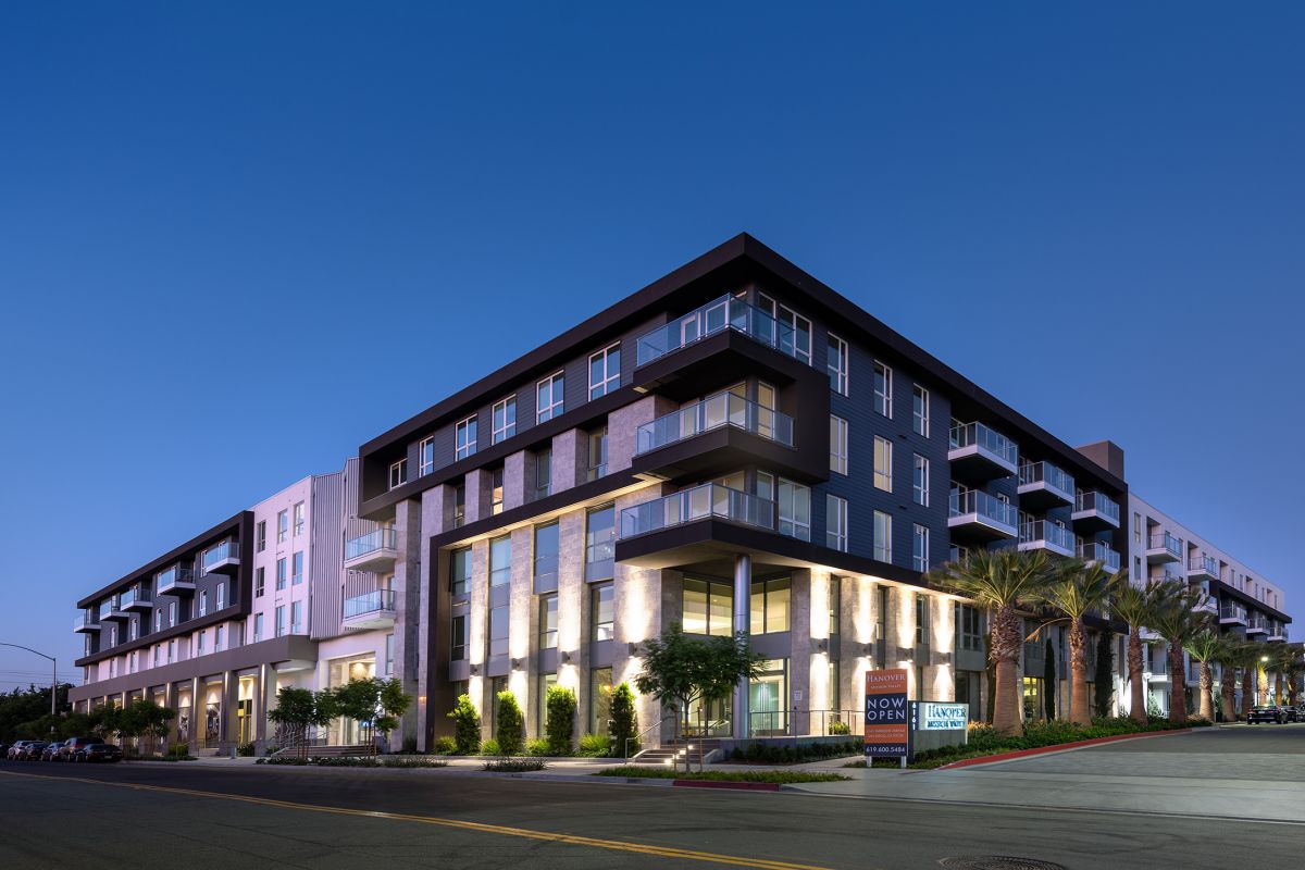 Modern apartment building at dusk with illuminated windows and palm trees along the street.