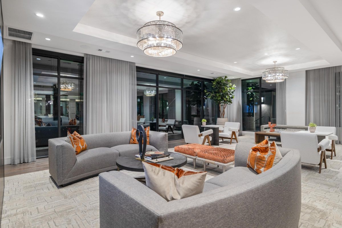 Modern lounge with curved gray sofas, orange pillows, and chandeliers. Large windows and indoor plants add elegance.