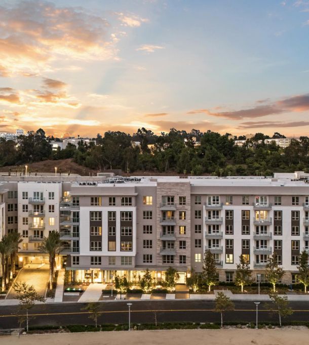 Modern apartment complex at sunset with a backdrop of trees and a colorful sky.